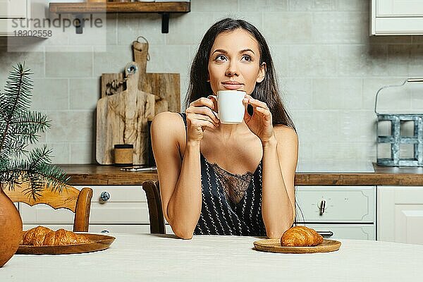 Fröhliche gemischtrassige junge Frau  die morgens in der Küche frischen Kaffee trinkt