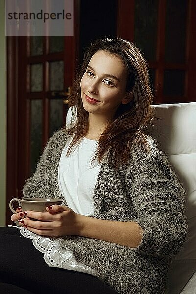 Glückliches lächelndes Mädchen mit großer Tasse Tee in den Händen auf einem Sessel im Zimmer