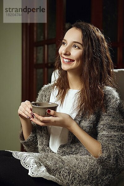Glückliches lächelndes Mädchen mit großer Tasse Tee in den Händen auf einem Sessel im Zimmer
