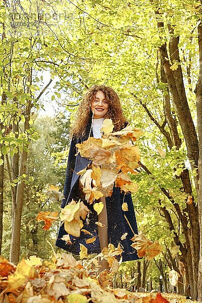 Fröhliche junge Frau tritt mit dem Fuß gegen Herbstblätter