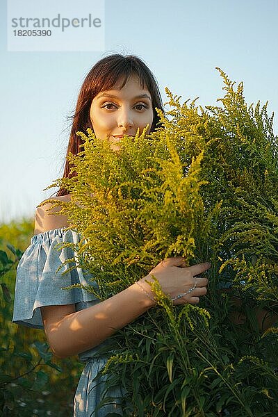 Lustiges Mädchen in himmelblauem Kleid mit Strauß Feldblumen im Sonnenuntergang