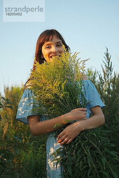 Lustiges Mädchen in himmelblauem Kleid mit Strauß Feldblumen im Sonnenuntergang