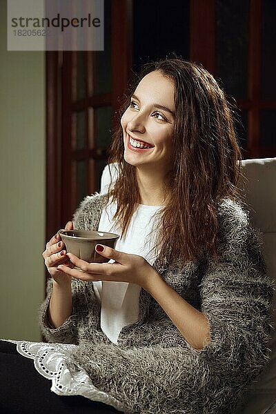 Glückliches lächelndes Mädchen mit großer Tasse Tee in den Händen auf einem Sessel im Zimmer