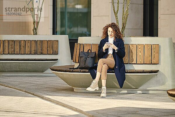 Ernste Frau  die auf einer Bank sitzt und Nachrichten auf ihrem Smartphone im Freien überprüft