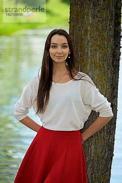 Porträt eines Mädchens in rotem Rock und weißer Bluse  das an einem Baum am Flussufer lehnt
