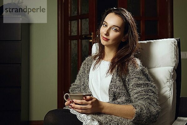 Glückliches lächelndes Mädchen mit großer Tasse Tee in den Händen auf einem Sessel im Zimmer