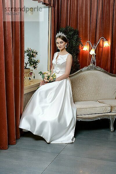 Schöne Braut in lond Kleid sitzt auf der Couch am Fenster