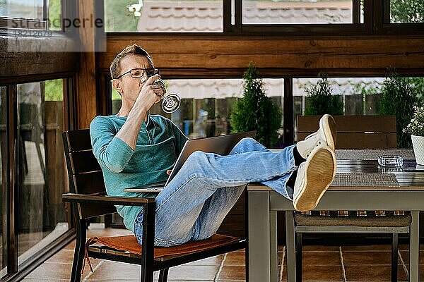 Ein Mann mittleren Alters trinkt Wein  während er auf der Veranda seines Landhauses eine Online-Sendung auf seinem Laptop verfolgt