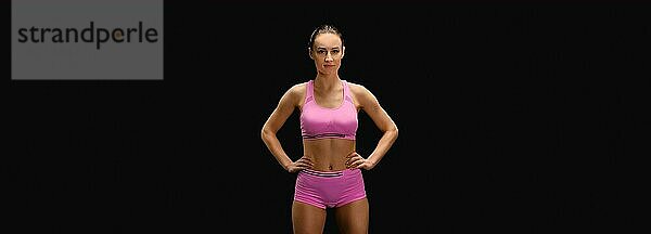 Sportliche junge Frau in rosafarbener Sportkleidung mit Händen auf der Hüfte vor schwarzem Hintergrund