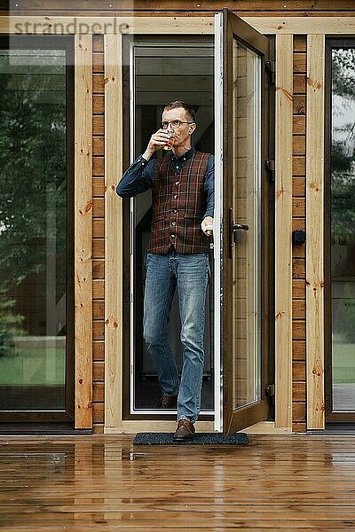 Ein eleganter Mann mittleren Alters kommt mit einem Glas in der Hand auf die Terrasse eines kleinen Holzhauses