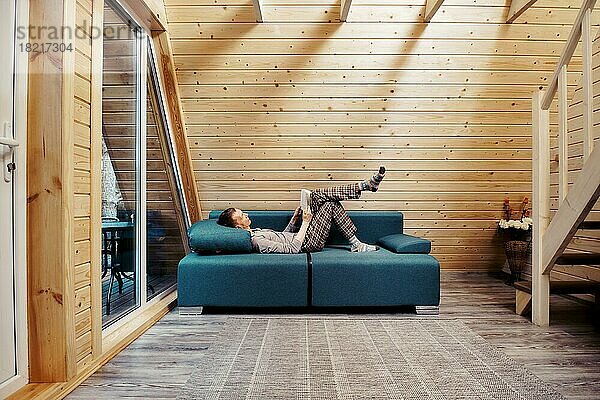 Mann mittleren Alters entspannt sich mit einem Buch auf dem Sofa in einem Chalet. Mann in karierten Hosen  langen Ärmeln und Wollsocken genießt die Ruhe auf dem Lande
