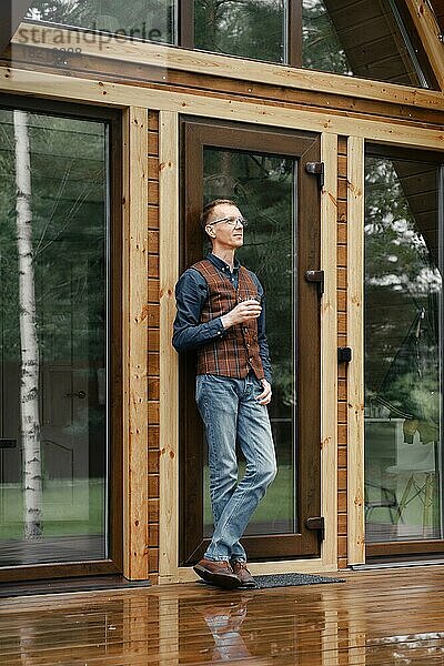 Nachdenklicher Mann mittleren Alters steht auf der Terrasse eines Holzbungalows und trinkt Bourbon