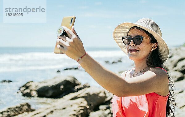 Mädchen mit Hut am Strand macht ein Selfie. Junge Frau im Urlaub  die Fotos am Strand macht  Lächelndes Mädchen mit Hut  das Fotos am Strand macht. Strandurlaub Konzept