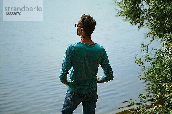 Rückansicht eines Mannes  der am Ufer eines Sees steht und auf das Wasser schaut