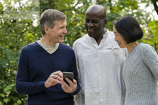 Porträt von drei Freunden  die beim Abhängen im Freien im öffentlichen Park auf den Bildschirm ihres Mobiltelefons schauen
