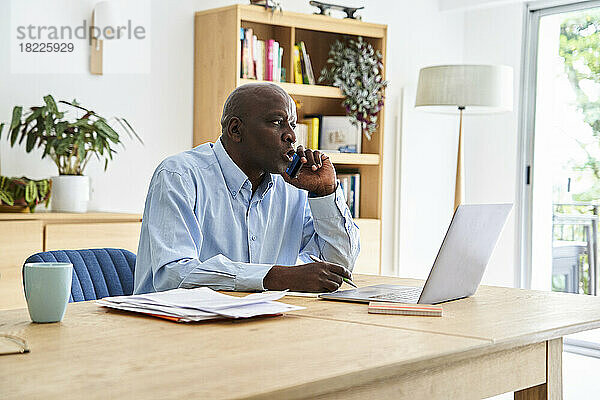 Afroamerikanischer Mann mittleren Alters telefoniert  während er zu Hause mit einem Laptop arbeitet