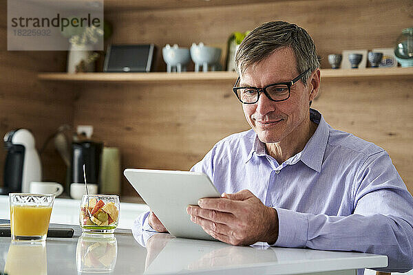 Ein professioneller Mann mittleren Alters lächelt und überprüft Nachrichten auf einem digitalen Tablet  während er zu Hause auf der Küchentheke frühstückt