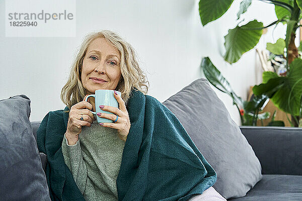 Ältere Frau sitzt auf der Couch und hält eine Tasse Tee in der Hand
