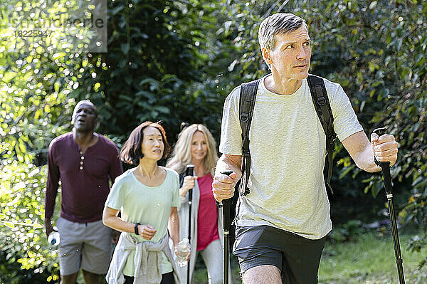 Middle-aged man carrying backpack and hiking poles while hiking in the woods with diverse group of friends