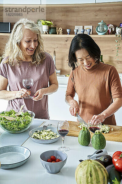 Ältere asiatische Frau schneidet Gemüse  während sie mit einer Freundin Wein trinkt