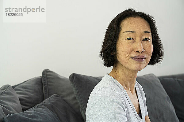 Porträt einer asiatisch-amerikanischen Seniorin  die in die Kamera blickt und im Wohnzimmer sitzt