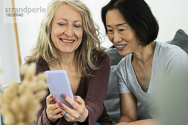 Portrait of two middle age female friends looking a mobile phone screen inside living room