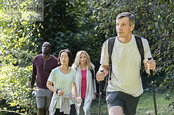 Middle-aged man carrying backpack and hiking poles while hiking in the woods with diverse group of friends