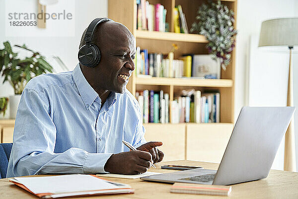 Ein afroamerikanischer Mann mittleren Alters trägt Kopfhörer und macht sich Notizen  während er an einer Fernkonferenz teilnimmt  während er zu Hause mit einem Laptop arbeitet