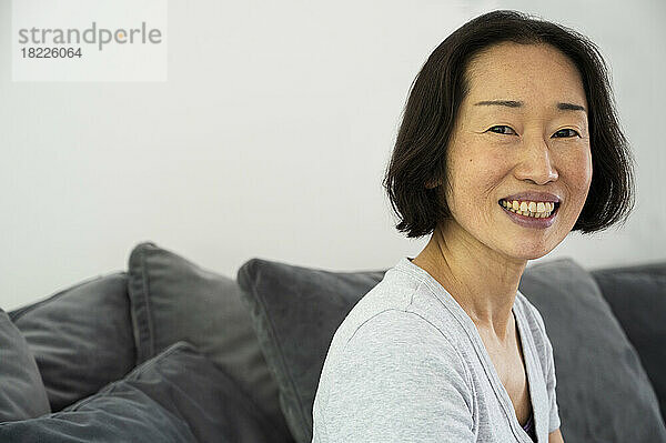 Porträt einer asiatisch-amerikanischen Seniorin  die lächelt und in die Kamera schaut  während sie im Wohnzimmer sitzt