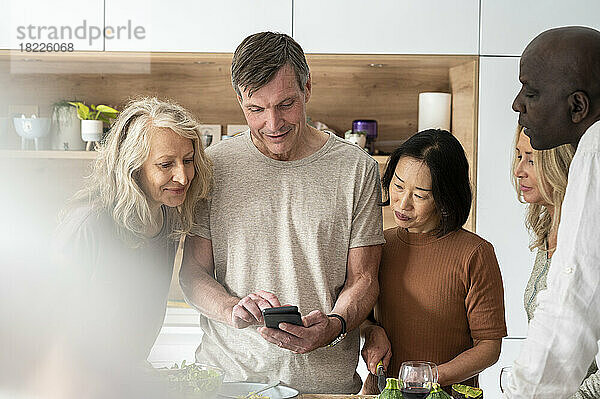 Eine bunt gemischte Gruppe älterer Freunde steht in der Küche und überprüft das Rezept auf dem Handy
