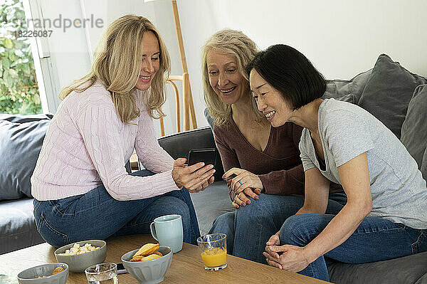 Porträt von drei Frauen mittleren Alters  die sich beim Betrachten von Fotos am Telefon im Wohnzimmer amüsieren