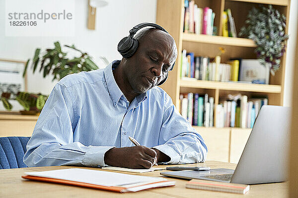 Reifer afroamerikanischer Mann  der sich bei einem Remote-Meeting mit Kopfhörern Notizen macht  während er zu Hause mit einem Laptop-Computer arbeitet