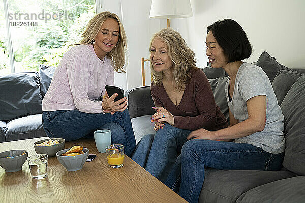 Halbbild einer Frau mittleren Alters  die zwei Freundinnen Fotos auf ihrem Handy zeigt  während sie zu Hause auf dem Sofa sitzt