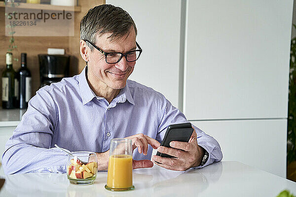 Ein professioneller Mann mittleren Alters lächelt und checkt Nachrichten am Telefon  während er zu Hause auf der Küchentheke frühstückt