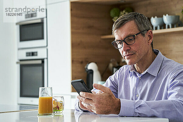 Berufstätiger Mann mittleren Alters checkt Nachrichten am Telefon  während er zu Hause auf der Küchentheke frühstückt