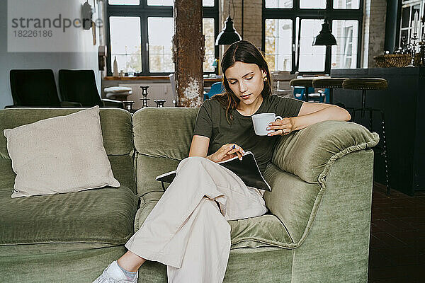 Junger Unternehmer liest ein Buch und trinkt einen Kaffee  während er auf dem Sofa im Studio sitzt