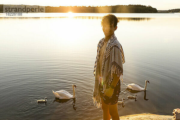 Porträt eines glücklichen Jungen  der in der Nähe eines Sees mit schwimmenden Schwänen im Hintergrund steht