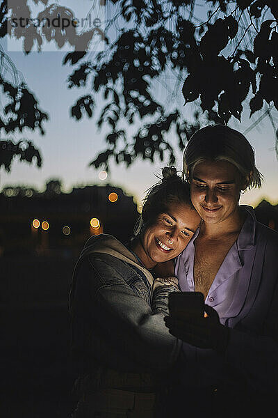 Lächelnde nicht-binäre Person  die nachts das Mobiltelefon einer Freundin benutzt