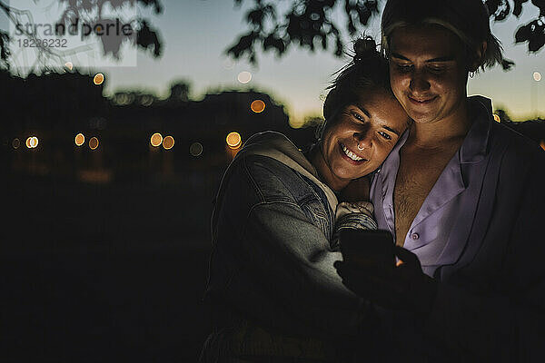 Lächelnde Frau umarmt nicht-binären Freund mit Handy in der Nacht