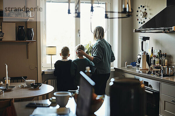 Rückansicht der Mutter mit Söhnen  die aus dem Küchenfenster nach draußen schauen