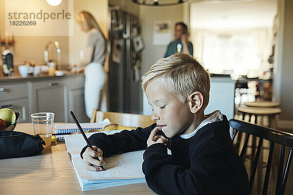 Nachdenklicher Junge macht Hausaufgaben in einem Buch  während er am Tisch sitzt