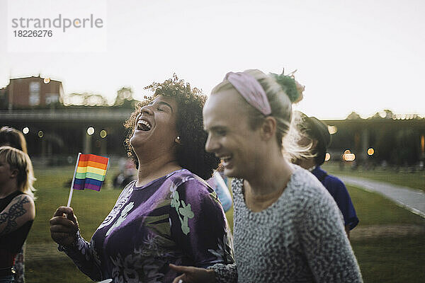 Fröhliche Transgender-Frau hält Regenbogenflagge  während sie sich mit einem nicht-binären Freund bei der Gay Pride Parade vergnügt
