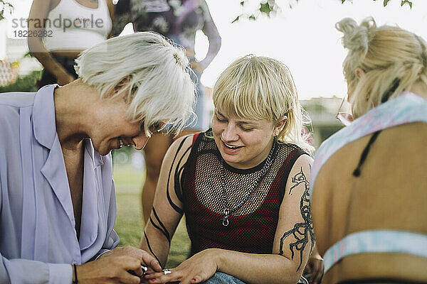 Lächelnde nicht-binäre Person trägt Nagellack auf einen Freund auf  der neben einer Frau sitzt