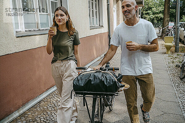 Männliche und weibliche Freunde trinken Kaffee  während sie mit dem Fahrrad auf dem Bürgersteig fahren