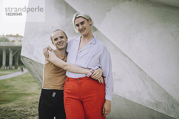 Porträt von lächelnden nicht-binären Freunden  die im Park an der Wand stehen