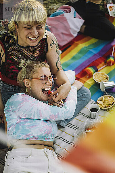 Glückliche nicht-binäre Person  die einer Freundin auf einer Picknickdecke liegend eine Erdbeere gibt