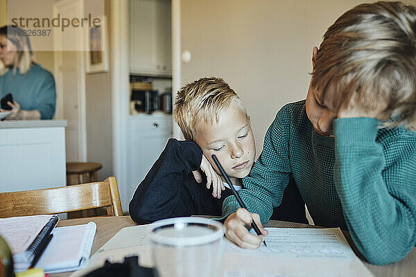 Junge stützt sich auf den Ellbogen  während er seinen Bruder bei den Hausaufgaben beobachtet