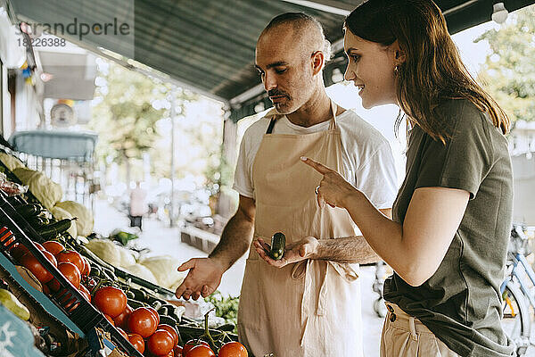 Seitenansicht einer Kundin  die mit einem männlichen Verkäufer spricht  der auf dem Markt Gemüse in der Hand hält
