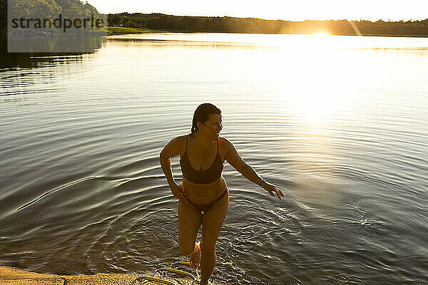 Glückliche Frau im Bikini geht nach dem Schwimmen bei Sonnenuntergang aus dem See