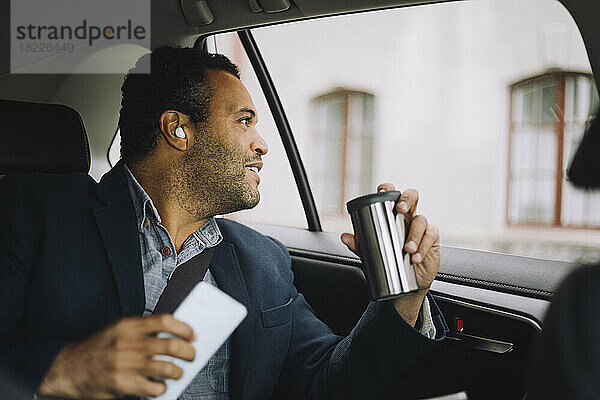 Lächelnder männlicher Pendler mit isoliertem Getränkebehälter und Smartphone  der aus dem Autofenster schaut